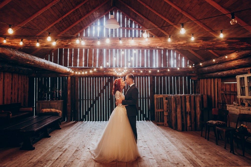 stylish groom and happy bride hugging under retro bulbs lights in wooden barn