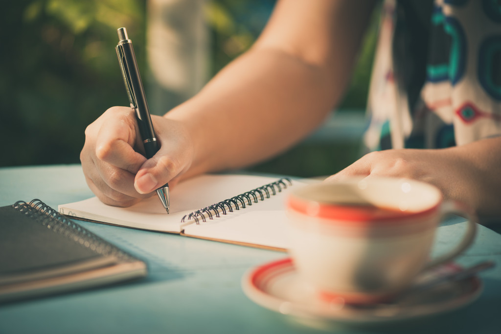 A person writing on a notebook while drinking coffee